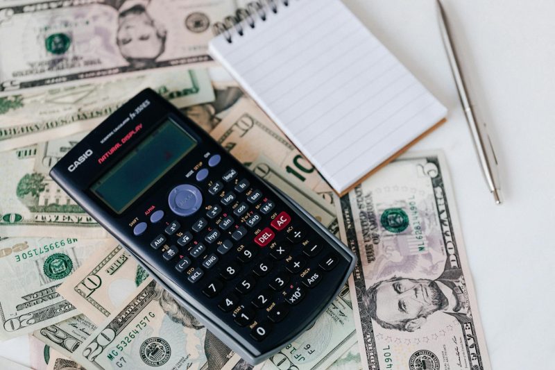 Electronic calculator and notepad atop United States dollar bills, accompanied by a metallic pen.