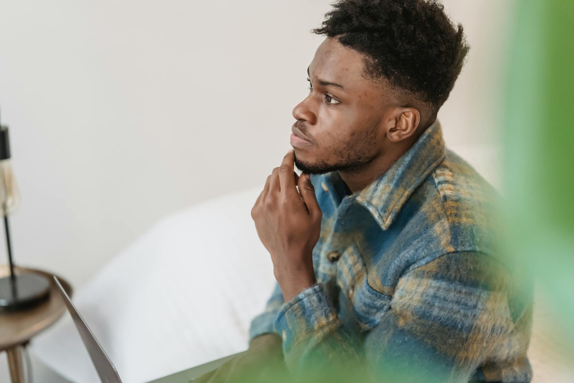 Young man sitting indoors, touching his chin thoughtfully in a tranquil, softly lit space.