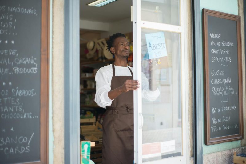 A man in an apron opens the door of a grocery store, welcoming customers in Portugal.