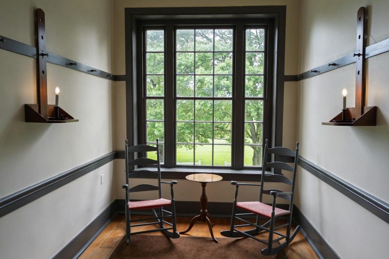 Cozy Shaker room with rocking chairs and candle sconces beside a bright window.