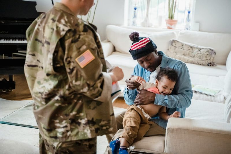 A smiling family, including a father and child, joyfully greets a military parent in a cozy home setting.